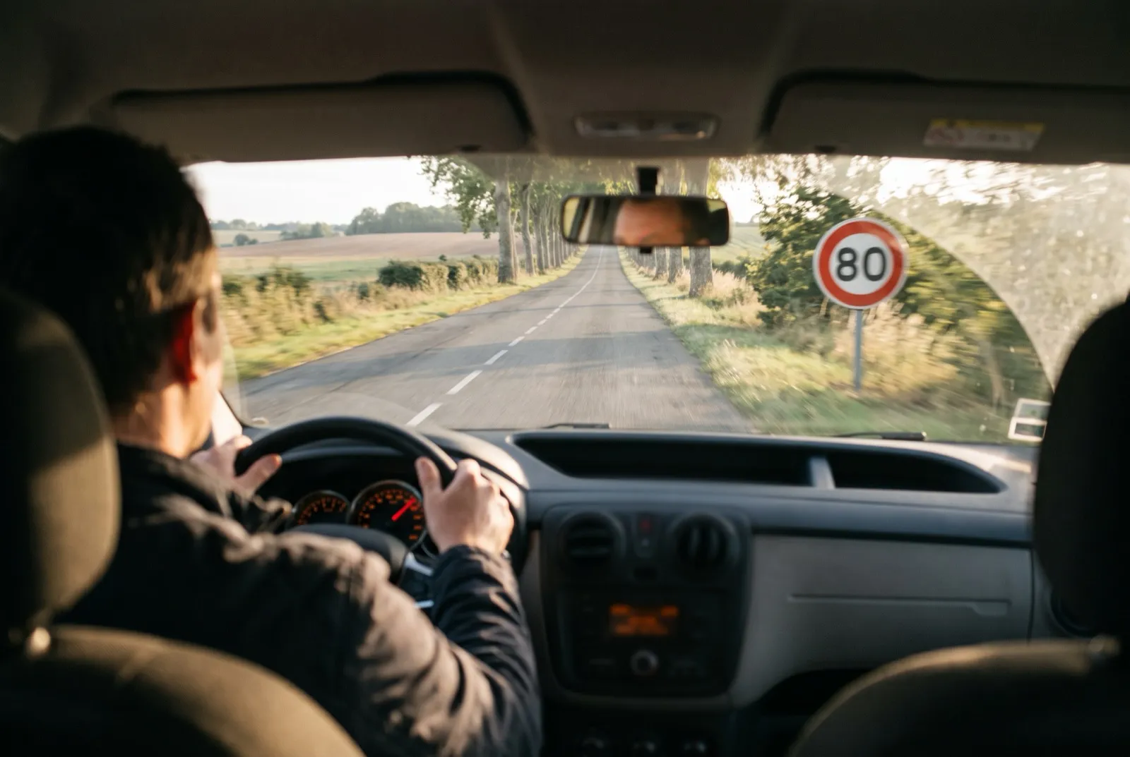 Conducteur vu de dos sur une route départementale bordée d’arbres, devant un panneau de limitation à 80 km/h